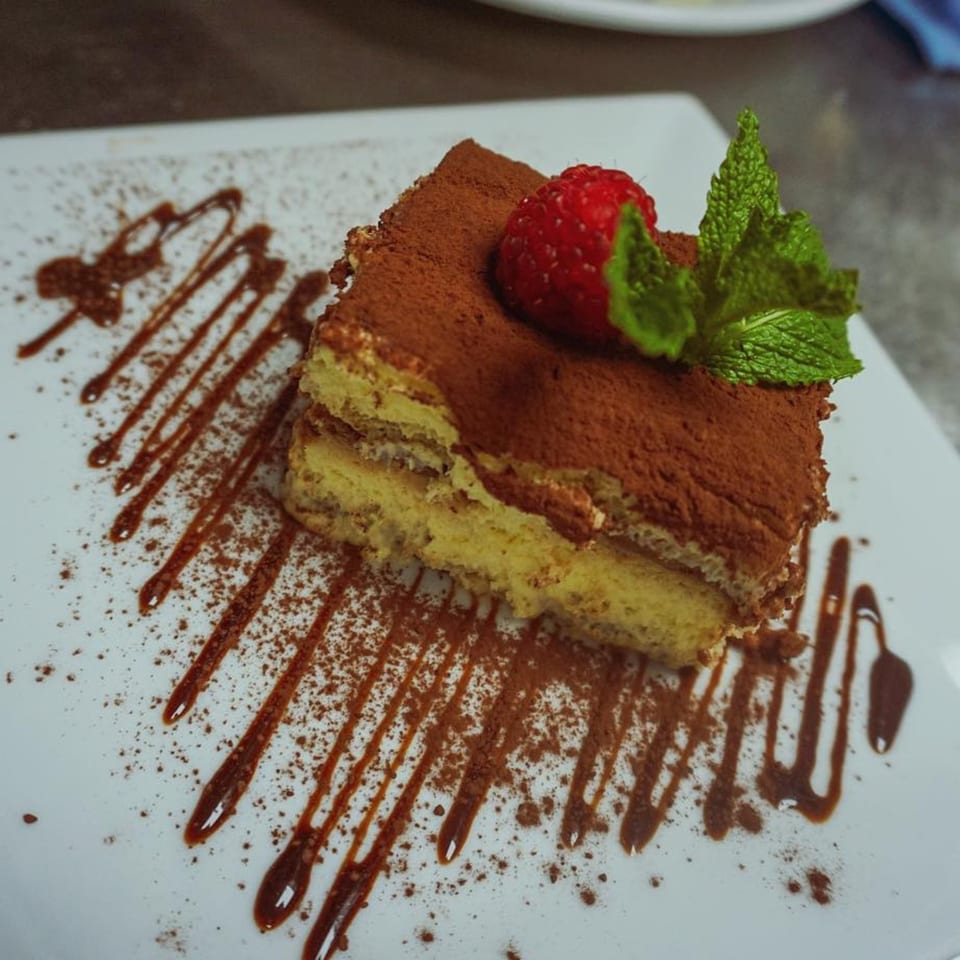 A slice of tiramisu with cocoa powder, a strawberry, and mint on a white plate with chocolate drizzle.