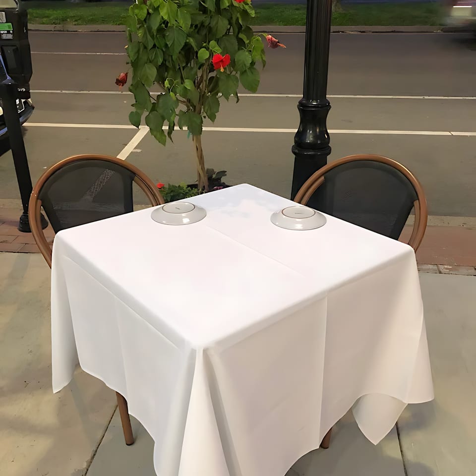 An outdoor dining table with a white tablecloth and two chairs, next to a potted plant.