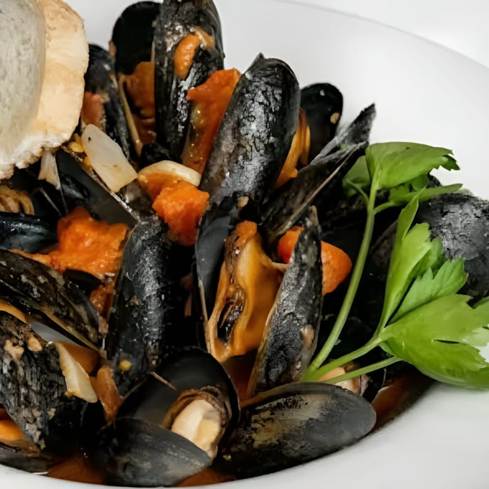 Steamed mussels in a rich orange sauce, garnished with parsley and a slice of bread.