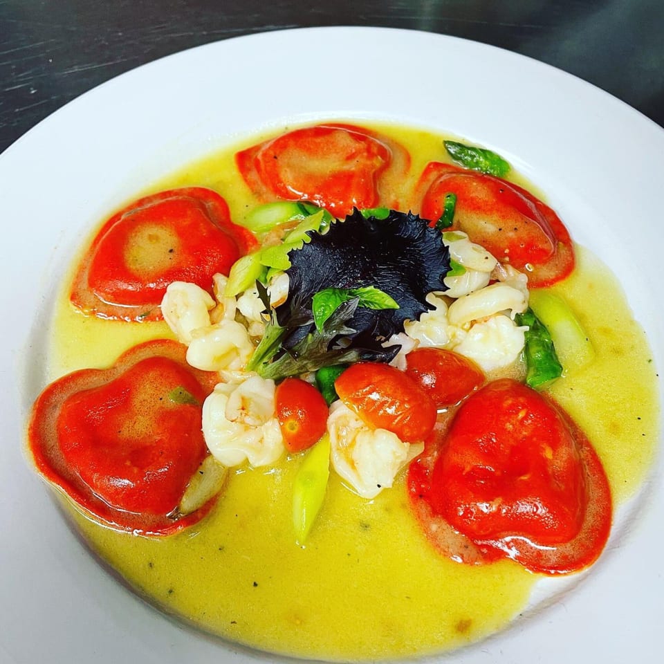 Heart-shaped red ravioli in a yellow sauce with shrimp, cherry tomatoes, and greens.