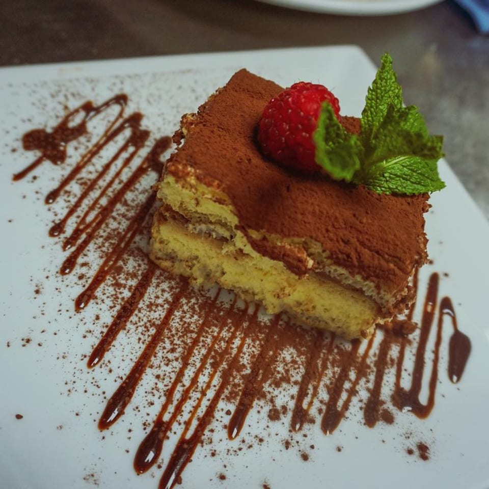 A slice of tiramisu with cocoa powder, a strawberry, and mint on a white plate with chocolate drizzle.