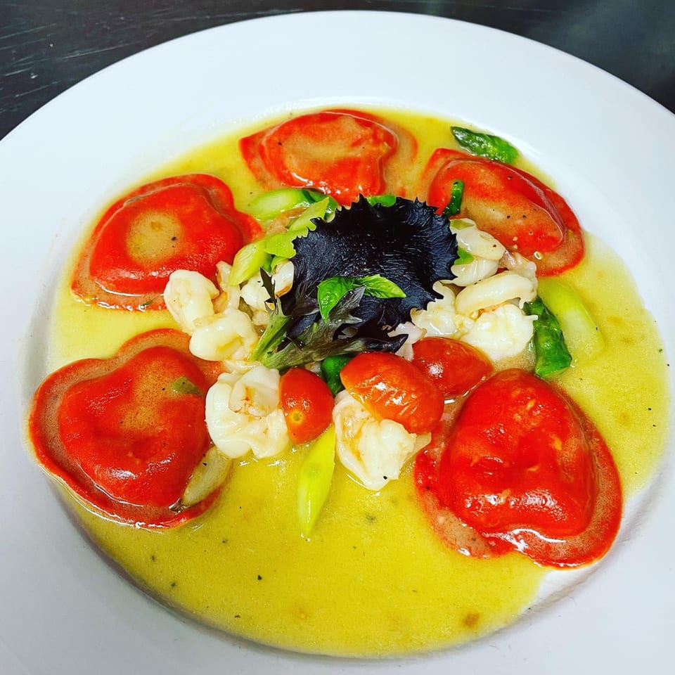 Red heart-shaped ravioli with shrimp, cherry tomatoes, asparagus, and greens in a yellow sauce.